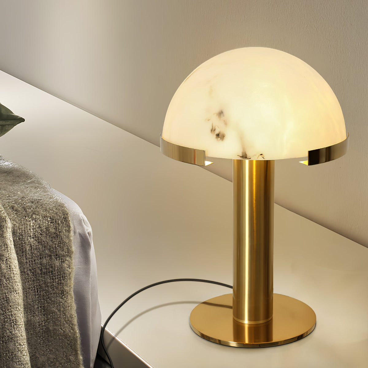 Lit Clérachic table lamp in gold, its alabaster dome glowing warmly on a pale shelf beside a knitted throw.