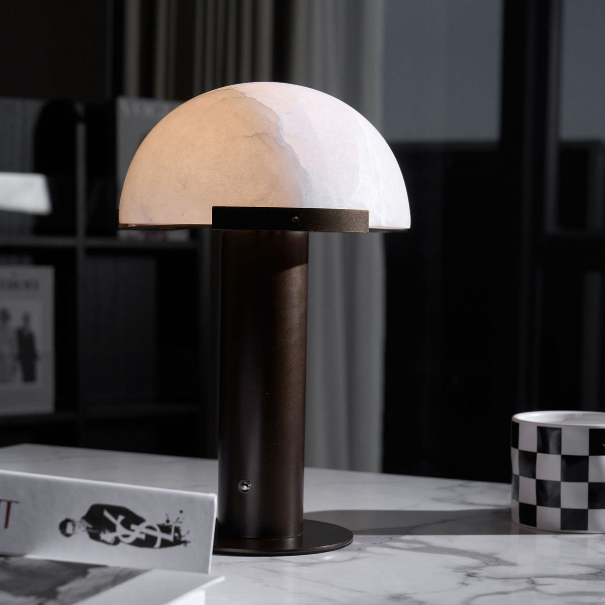 Dark bronze-coloured version of this Clérachic table lamp on a marble top, with a chequered mug and art books behind it.