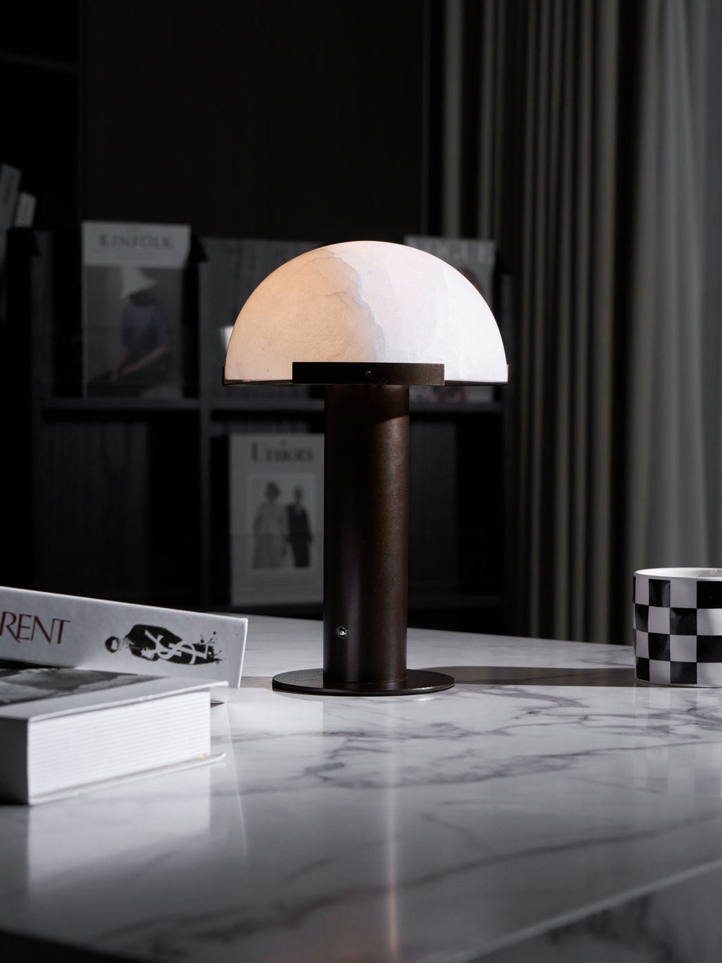 Narrow crop of the dark-based Clérachic table lamp on a white marble table, with dark shelving and grey curtains behind.