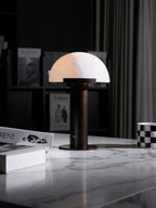 Narrow crop of the dark-based Clérachic table lamp on a white marble table, with dark shelving and grey curtains behind.