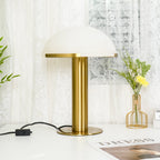 Gold-based Clérachic table lamp with a white dome, its black cord and inline switch trailing across a white desk beside a vase of yellow flowers.