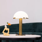 On a round marble-topped table, this Clérachic table lamp stands in gold in front of a deep green velvet sofa and a small figurine.