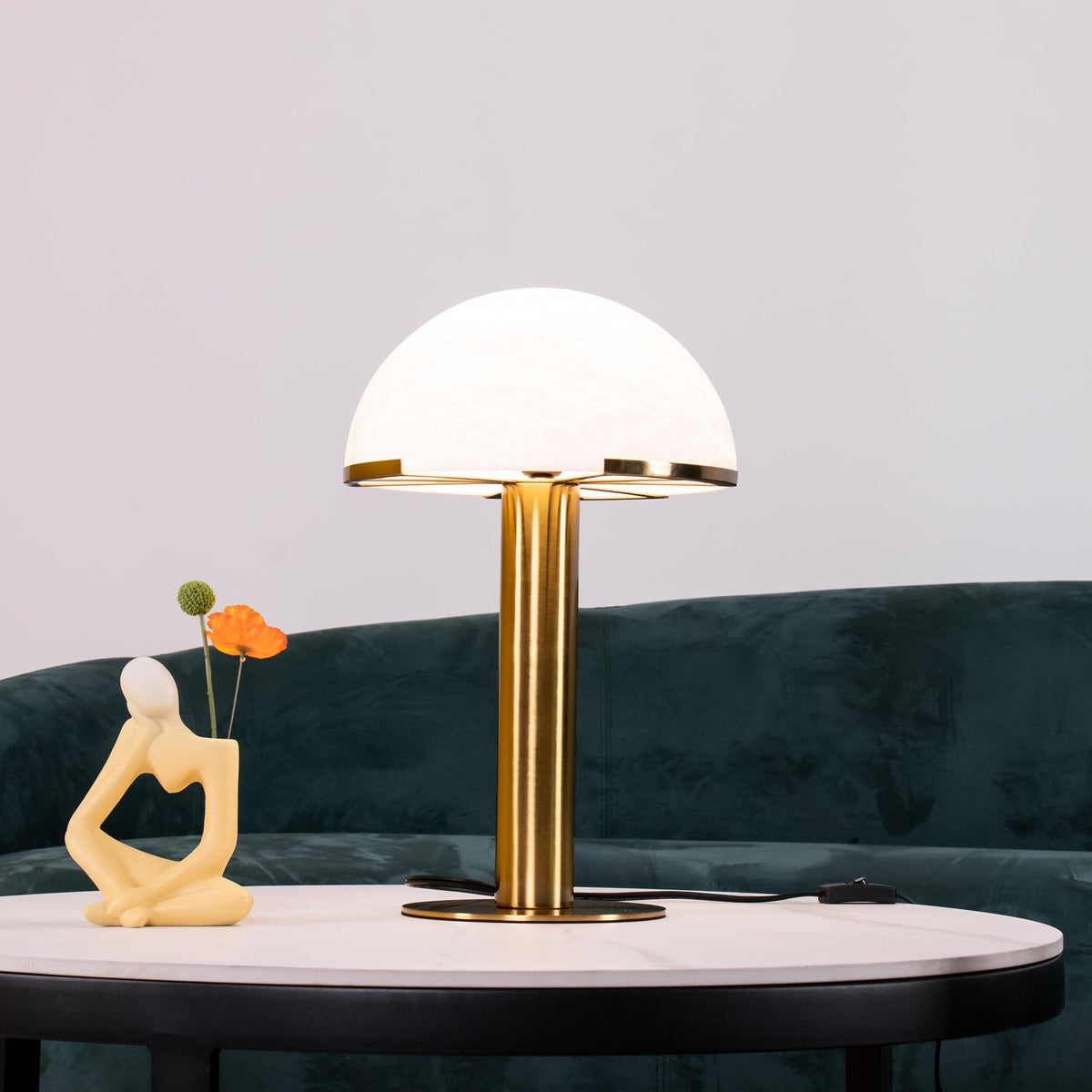 On a round marble-topped table, this Clérachic table lamp stands in gold in front of a deep green velvet sofa and a small figurine.