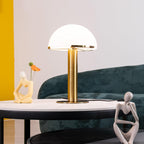 Gold Clérachic table lamp lit on a round tabletop, with a dark green sofa and a cream sculpture in the background.