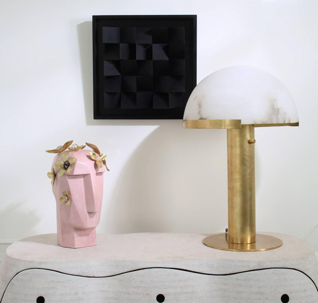 Gold Clérachic table lamp on a pale table, with a pink sculpted face vase and a black textured wall panel behind it.