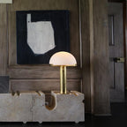 Softly lit Clérachic table lamp with a gold stem glowing against dark timber panelling, set on a block of pale travertine.