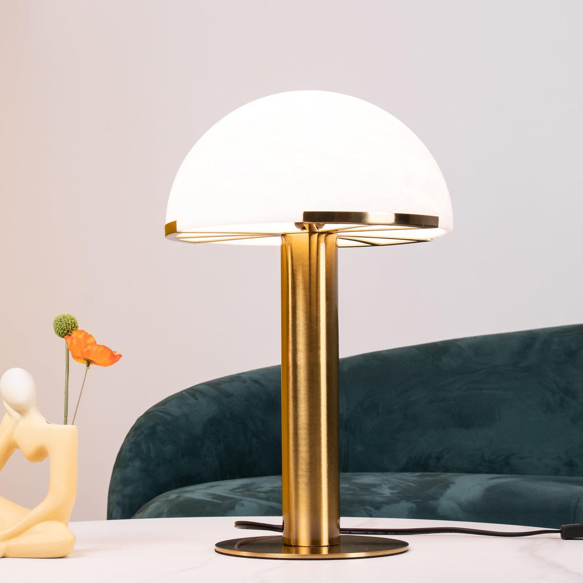 Close view of this Clérachic table lamp in gold, its frosted white dome lit from within, with a green sofa and cream figurine behind.