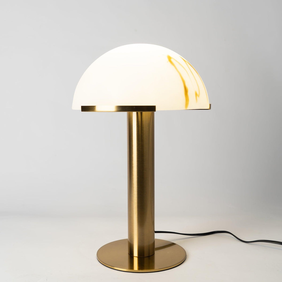 Gold Clérachic table lamp on a pale grey backdrop, its white dome streaked with amber veining and its black cord curling out from the base.