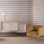 Gold Clérachic table lamp on a cream cabinet in a room with striped neutral wallpaper and an ochre velvet armchair beside it.