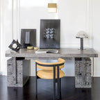 Dark-based Clérachic table lamp on a weathered grey oak desk, with framed black artworks and a tan leather chair in a white room.