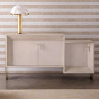 Gold Clérachic table lamp on top of a pale cabinet with brass handles, set against horizontally striped wallpaper.