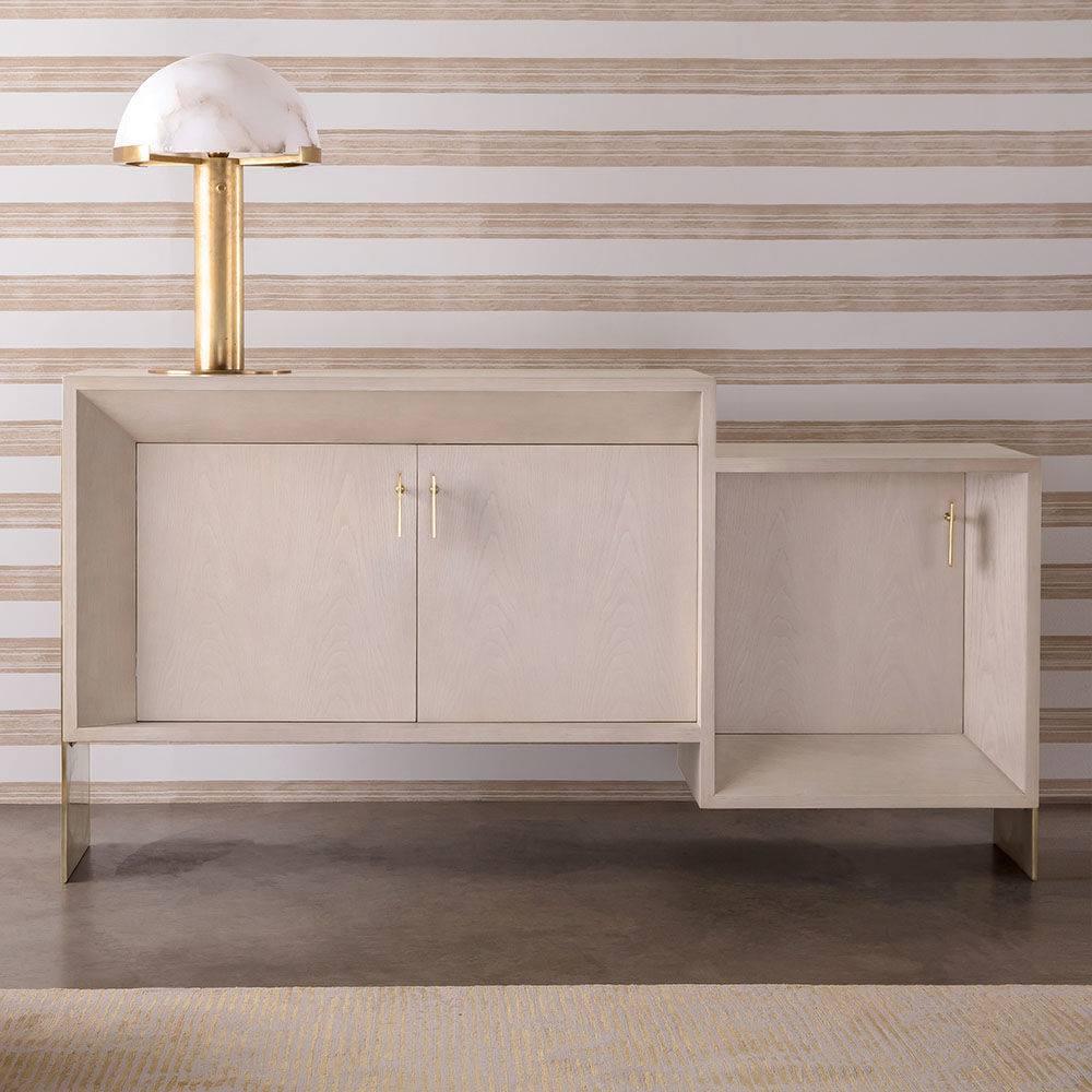 Gold Clérachic table lamp on top of a pale cabinet with brass handles, set against horizontally striped wallpaper.