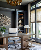 Gold Clérachic table lamp on a wooden desk in a dark navy study, with built-in shelves and bamboo blinds at the windows.