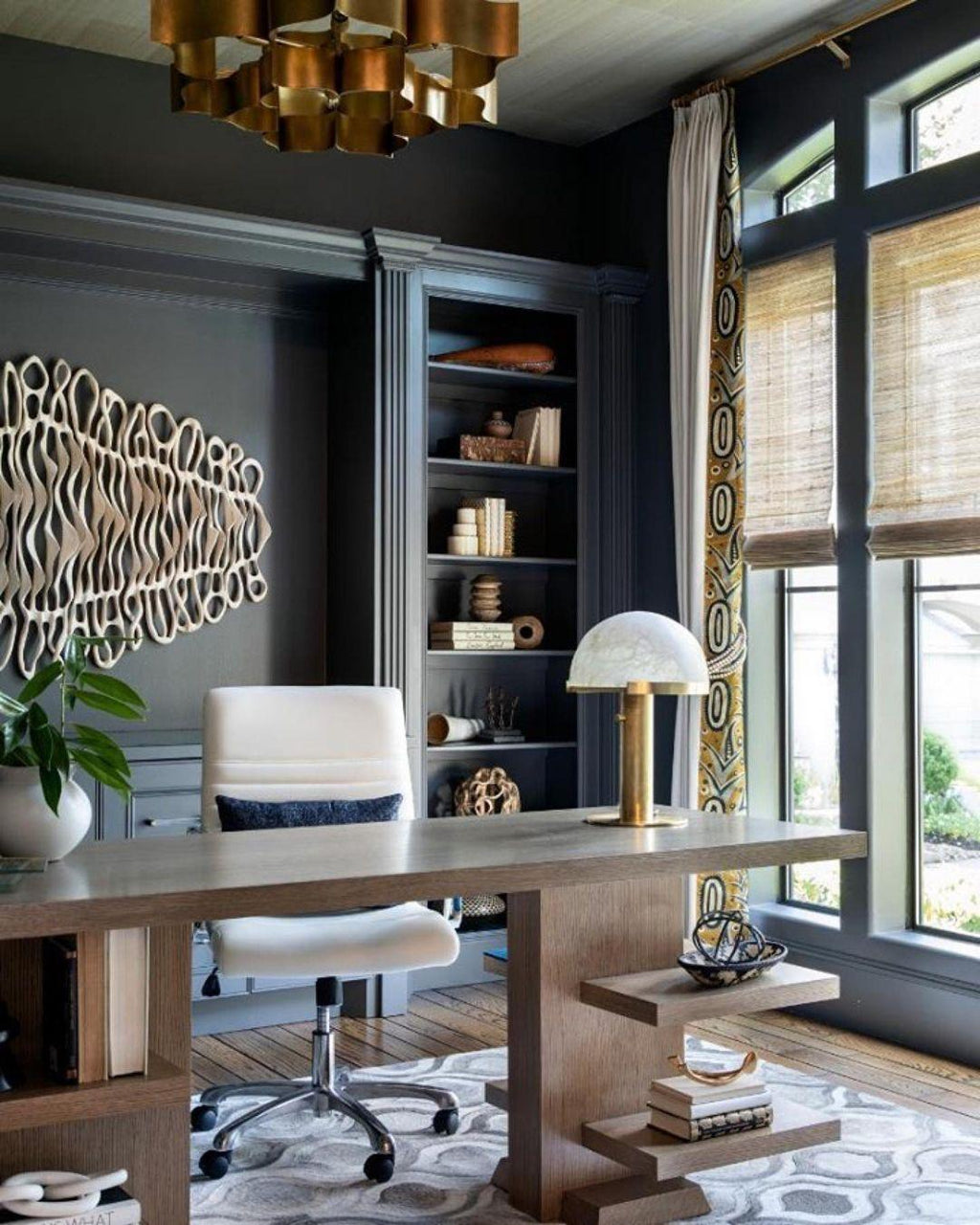 Study scene with a Clérachic table lamp in gold on a wooden desk, a white office chair beside it and a brass ceiling fitting overhead.