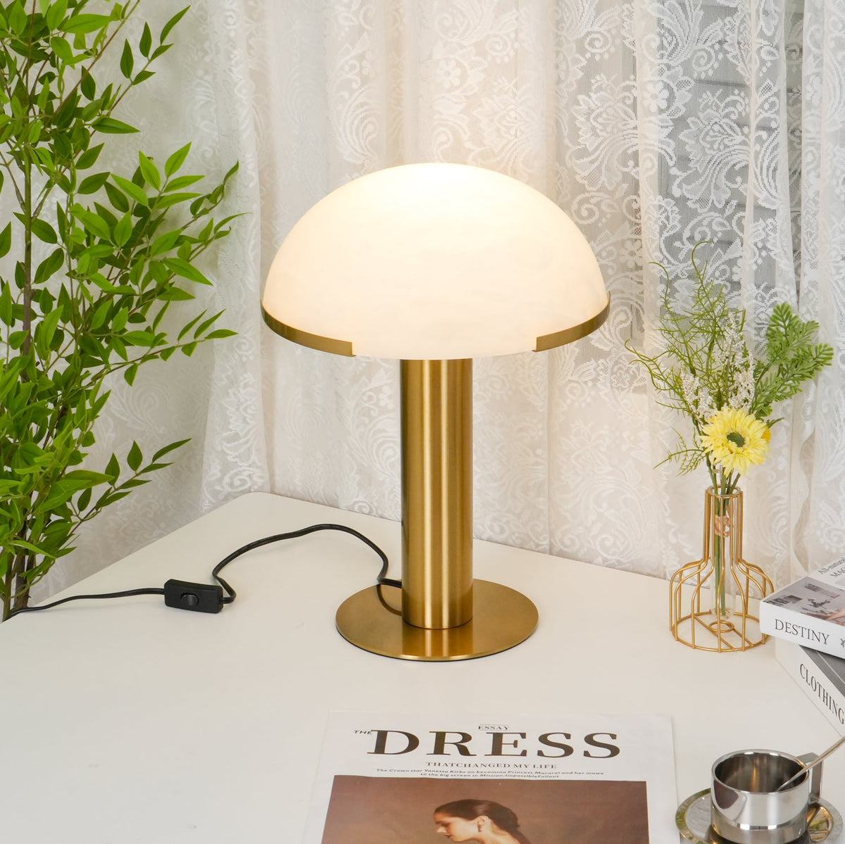 Gold Clérachic table lamp on a white desk by a lace curtain, with a leafy plant, a vase of yellow flowers and a magazine nearby.