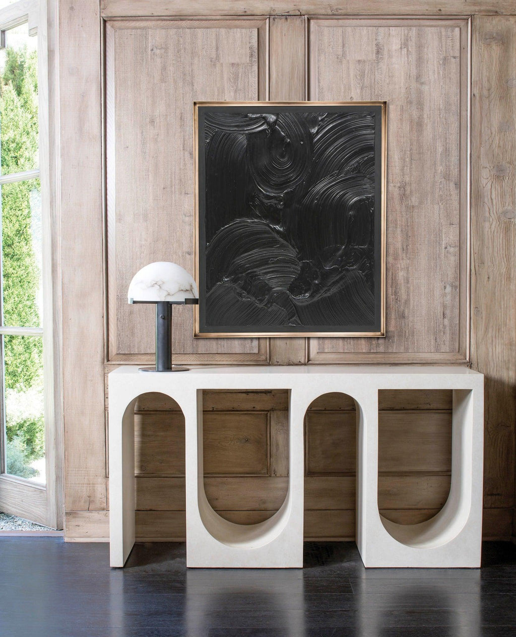 Dark-based Clérachic table lamp on a white arched console, below a large black textured painting on a pale timber wall.
