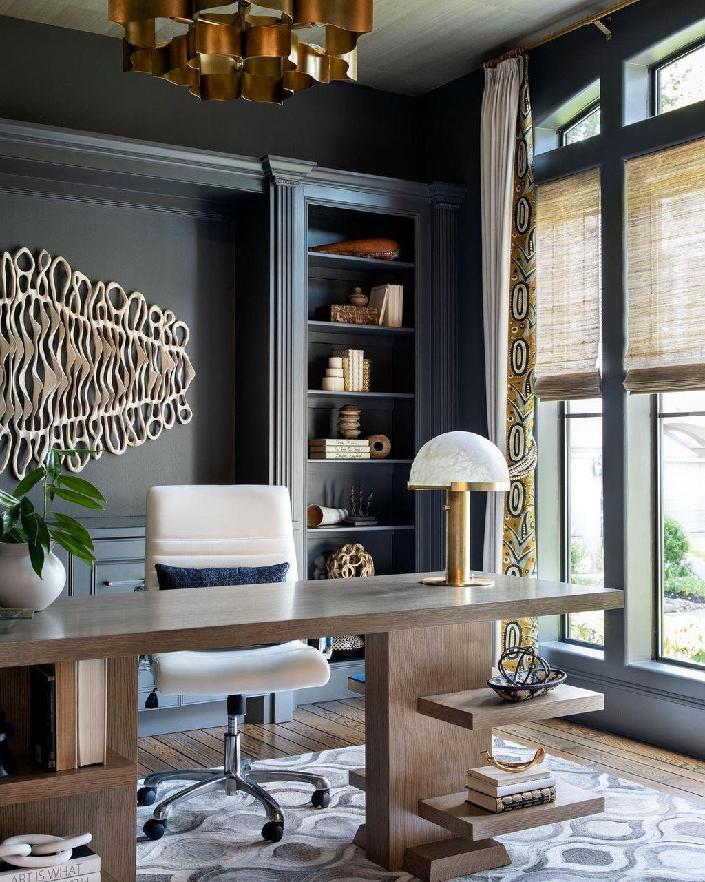 Gold Clérachic table lamp beside a white office chair in a study with dark joinery, a patterned rug and bamboo blinds.