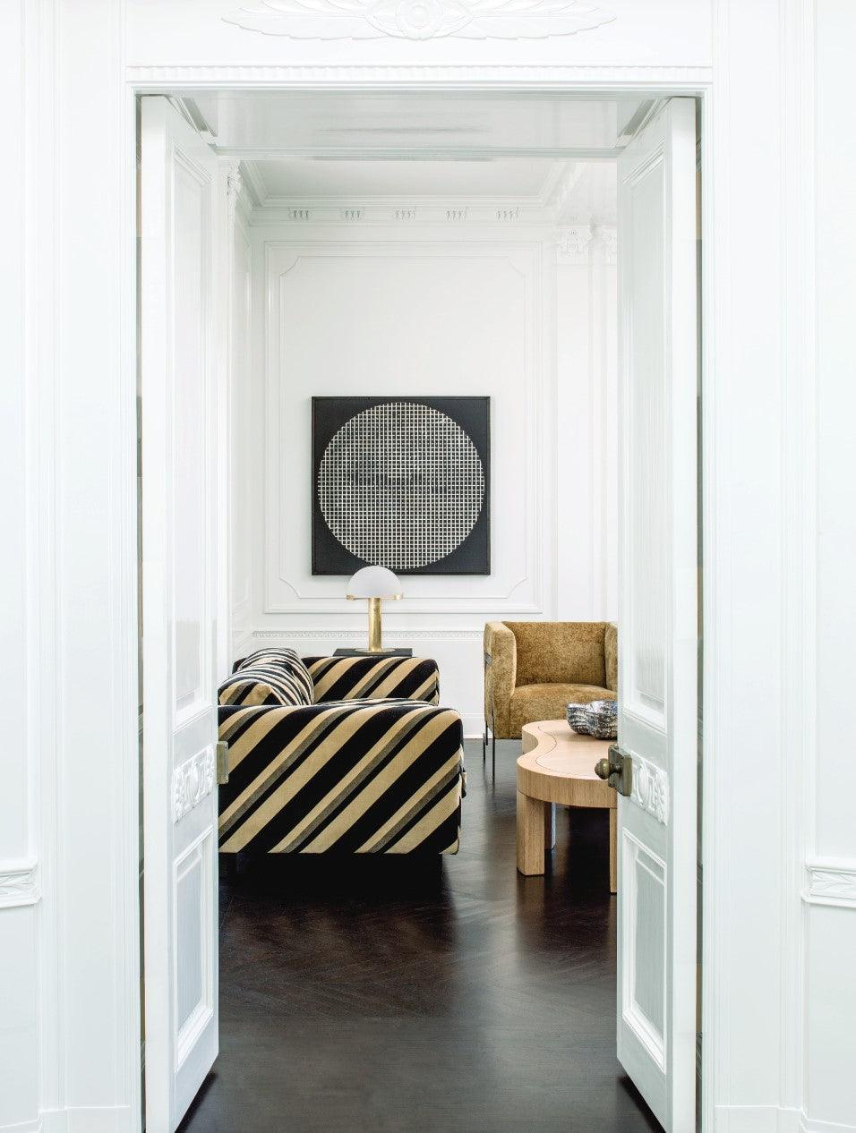 Distant view through a white doorway, with a small Clérachic table lamp in gold on a table in a bright room with striped seating.