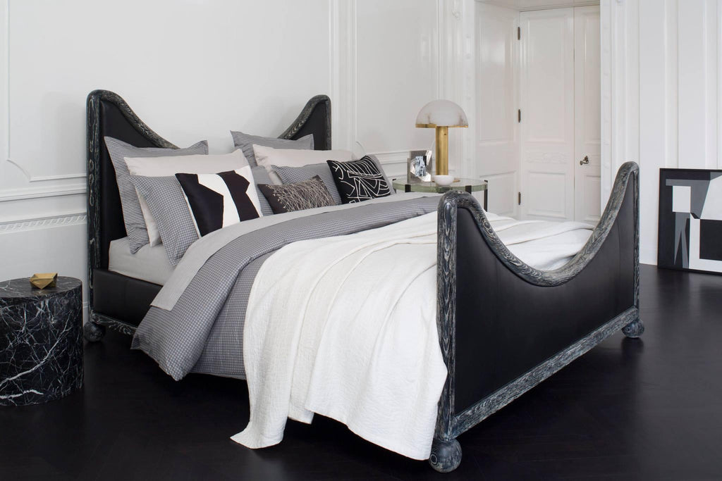 Gold Clérachic table lamp on a bedside table next to a black sculptural bed dressed in white and grey linen.