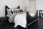 Gold Clérachic table lamp on a bedside table next to a black sculptural bed dressed in white and grey linen.