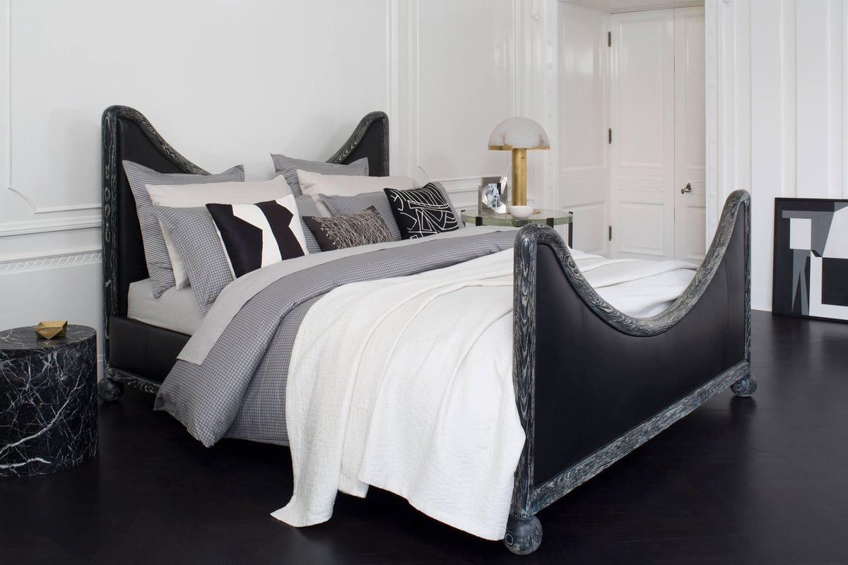 Gold Clérachic table lamp on a bedside table next to a black sculptural bed dressed in white and grey linen.