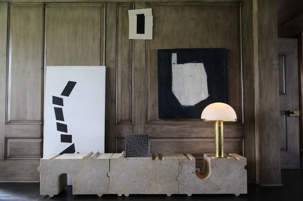Gold Clérachic table lamp with a pale dome on a block of cream stone, in front of dark timber panelling and abstract paintings.