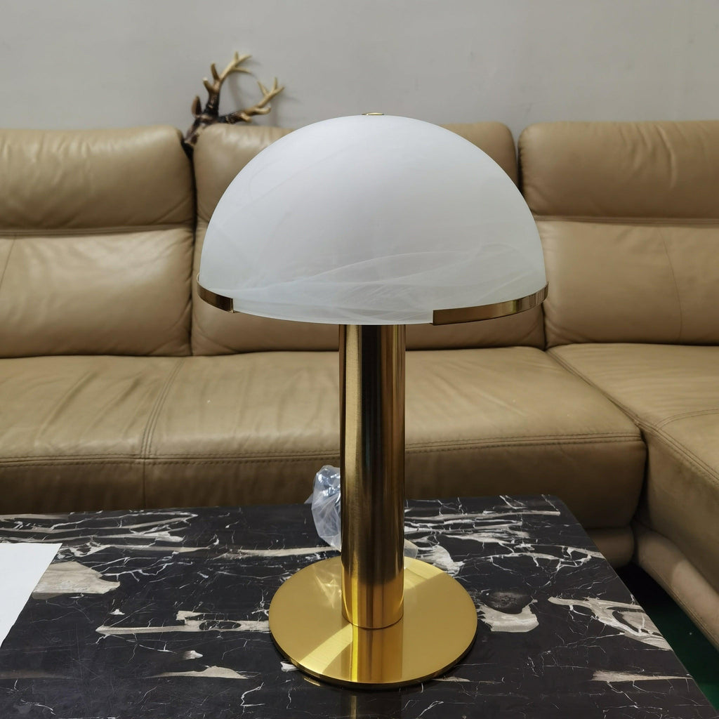 Gold Clérachic table lamp with a smooth frosted white dome on a black marble table, in front of a tan leather sofa.