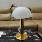 Gold Clérachic table lamp with a smooth frosted white dome on a black marble table, in front of a tan leather sofa.