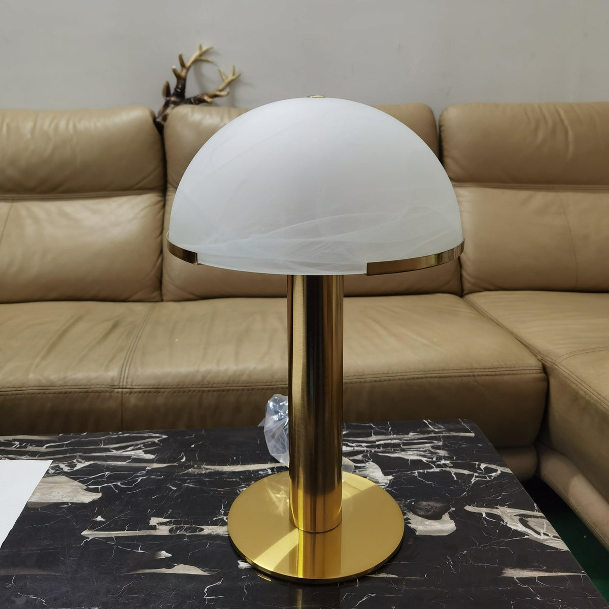 Gold Clérachic table lamp with a smooth frosted white dome on a black marble table, in front of a tan leather sofa.
