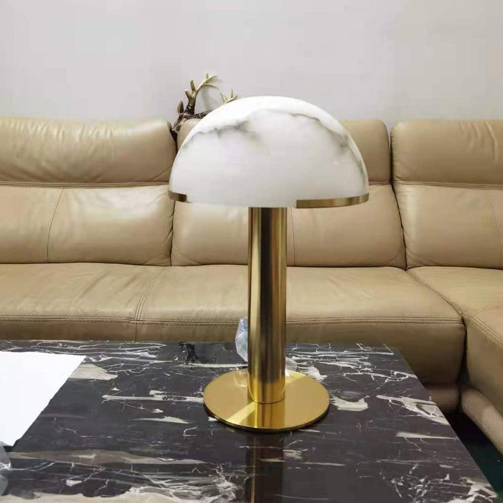 Same gold Clérachic table lamp with a white dome softly streaked in grey, on a black veined marble table before a leather sofa.