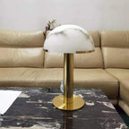 Same gold Clérachic table lamp with a white dome softly streaked in grey, on a black veined marble table before a leather sofa.