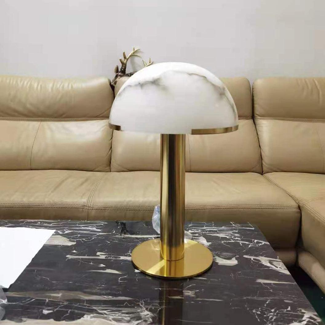 Same gold Clérachic table lamp with a white dome softly streaked in grey, on a black veined marble table before a leather sofa.