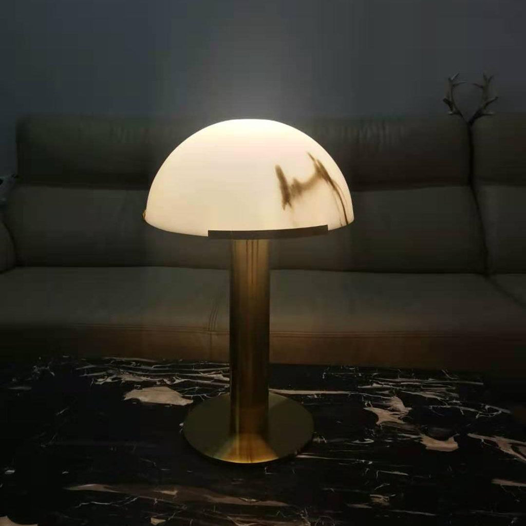 Gold Clérachic table lamp switched on in a dim living room, the dome giving off a warm glow over a dark marble table.