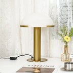 Gold Clérachic table lamp on a white table, with its cord and inline switch to the left and a vase of yellow flowers to the right.