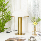 In front of a lace curtain, this gold Clérachic table lamp has a plain white dome, with a leafy plant and a magazine beside it.
