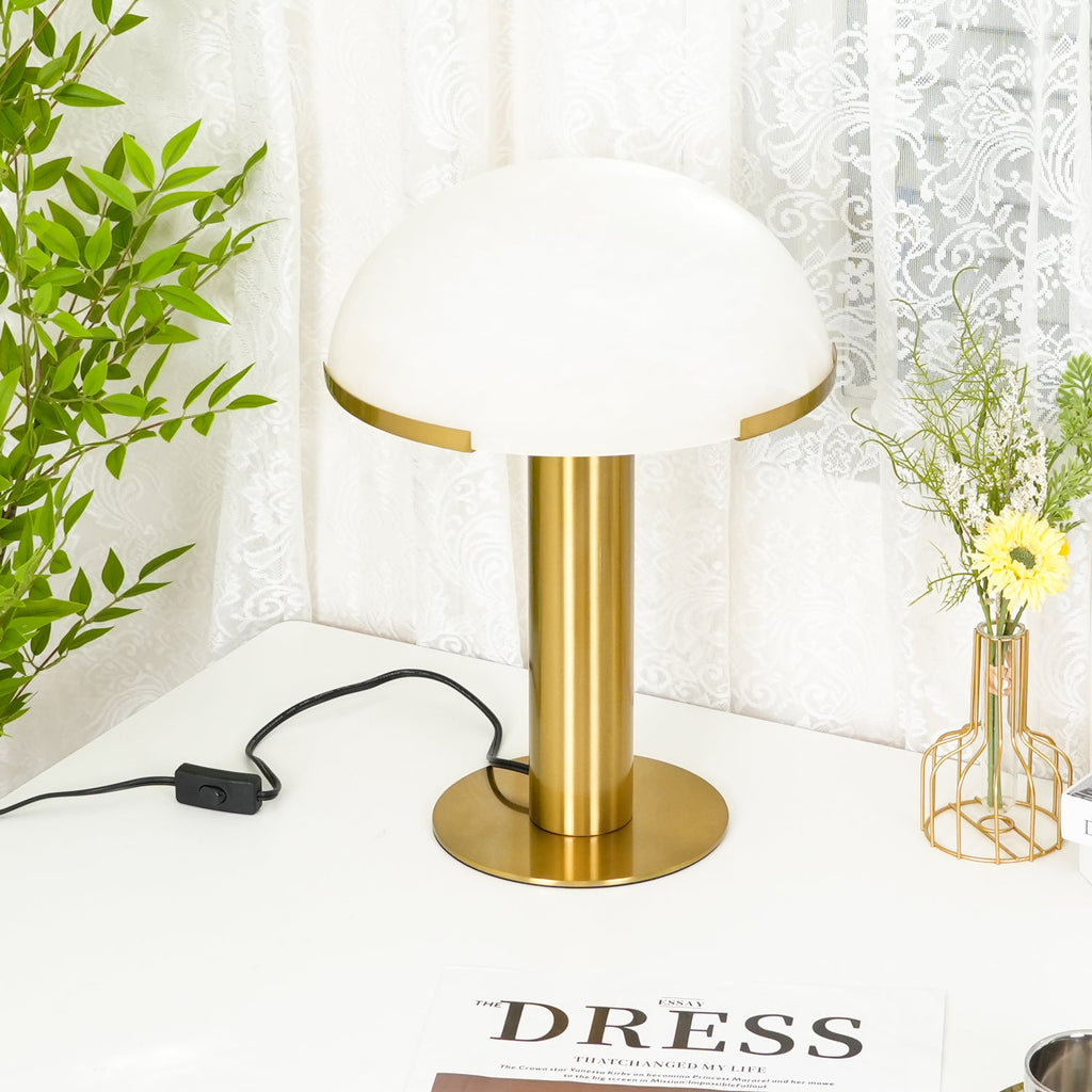 Gold Clérachic table lamp seen from a slightly raised angle, with the round base visible beneath a white dome and a plant to one side.