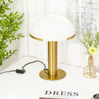 Gold Clérachic table lamp seen from a slightly raised angle, with the round base visible beneath a white dome and a plant to one side.