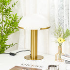 Gold Clérachic table lamp on a white tabletop, a small steel cup and a magazine beside the base and a yellow flower vase behind.
