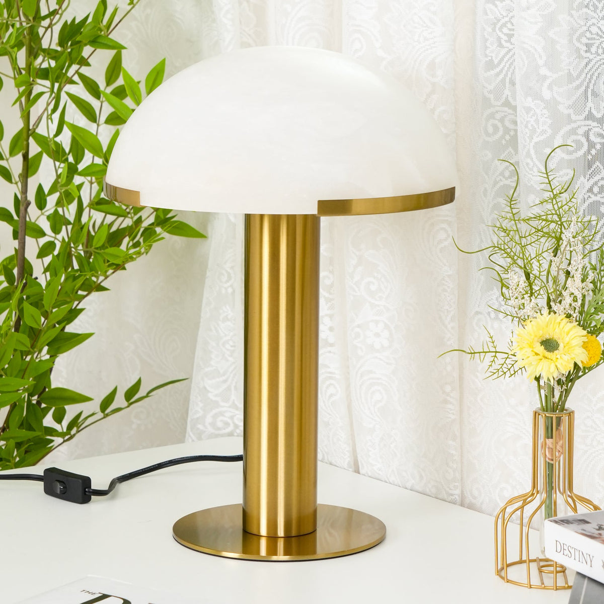 Larger view of this Clérachic table lamp in gold, with a white dome and a wide brass base next to a bunch of yellow flowers.