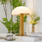 Gold Clérachic table lamp with its cord and switch on a desk, next to a steel cup on a saucer, books and a leafy plant.