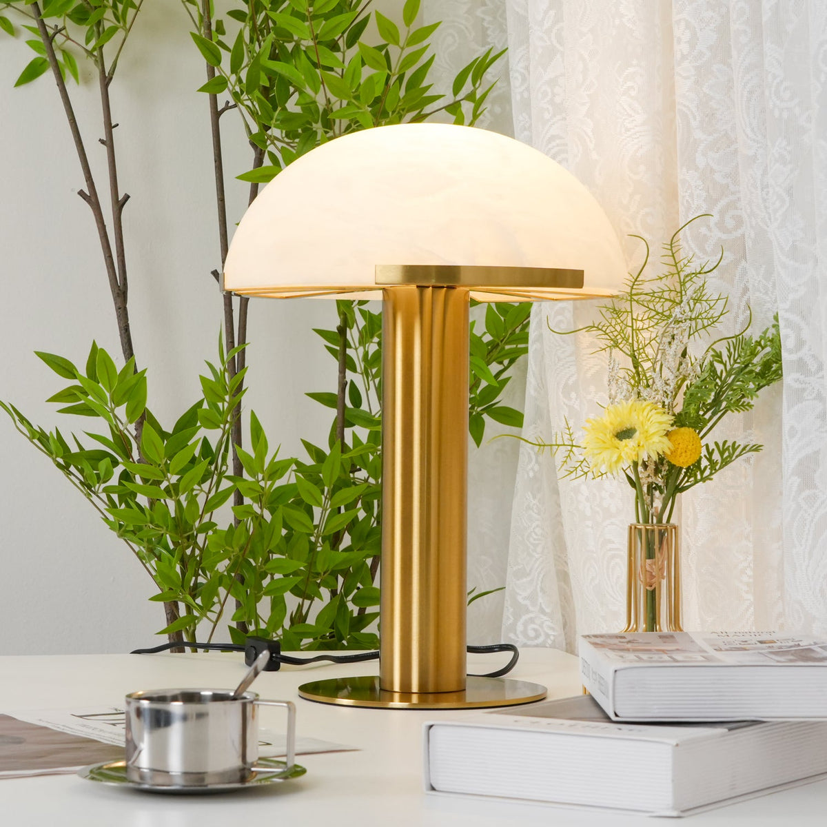 Gold Clérachic table lamp with its cord and switch on a desk, next to a steel cup on a saucer, books and a leafy plant.