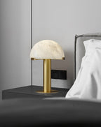 Gold Clérachic table lamp lit on a dark bedside shelf, with a grey padded headboard and white pillow beside it.