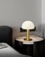 Gold Clérachic table lamp lit on a round dark bedside table, with a wall socket behind it and a grey bed at the side.