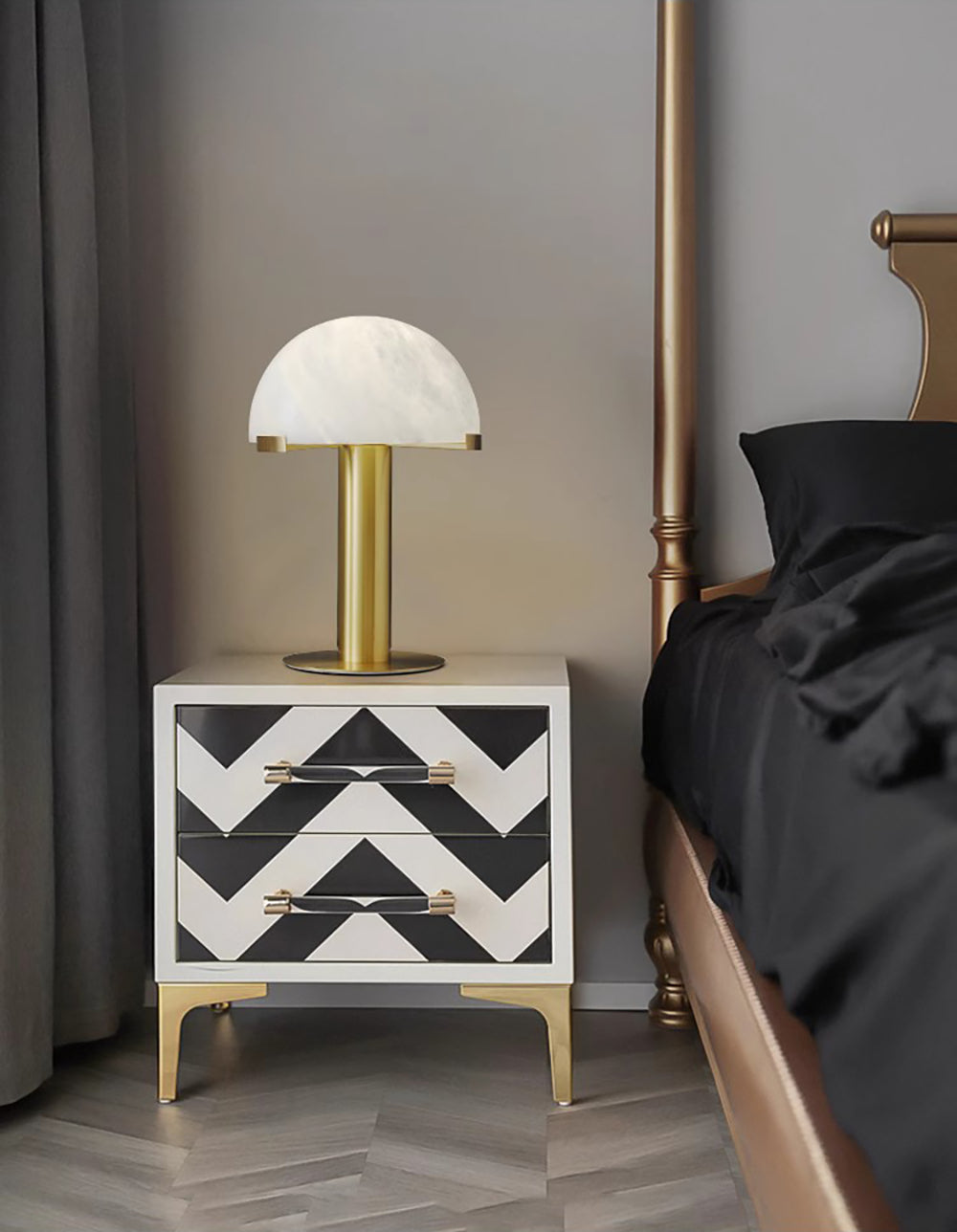 Gold Clérachic table lamp on a black and white chevron nightstand, next to a bed with dark linen and a brass four-poster.
