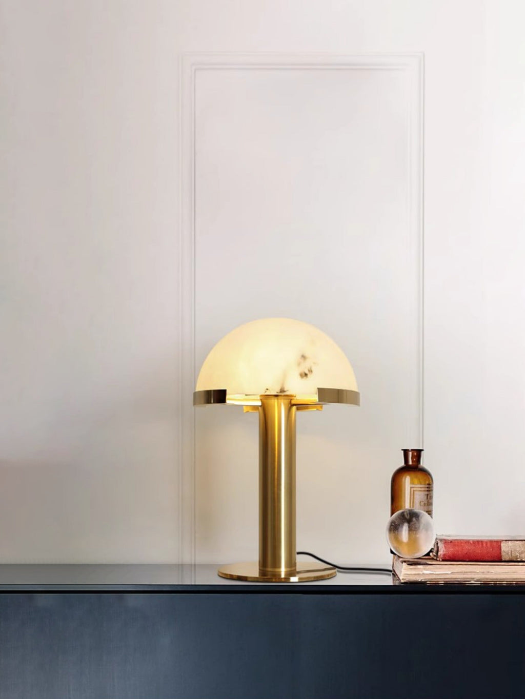 Gold Clérachic table lamp glowing on a dark blue console, with an amber glass bottle, a glass sphere and a stack of books beside it.