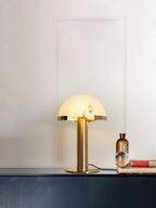 Gold Clérachic table lamp glowing on a dark blue console, with an amber glass bottle, a glass sphere and a stack of books beside it.