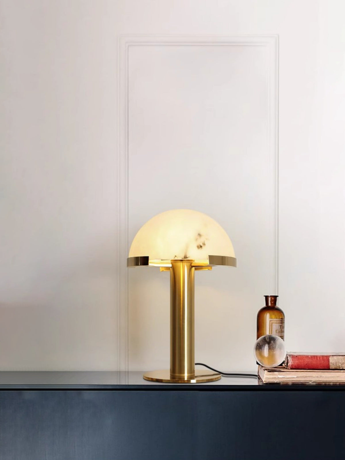 Gold Clérachic table lamp glowing on a dark blue console, with an amber glass bottle, a glass sphere and a stack of books beside it.