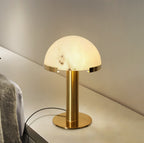 Gold Clérachic table lamp lit on a pale shelf, with a knitted blanket draped at the left and a cord looping behind.