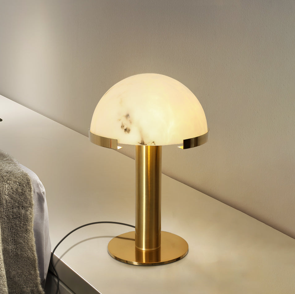 Gold Clérachic table lamp lit on a pale shelf, with a knitted blanket draped at the left and a cord looping behind.