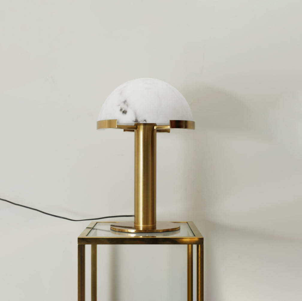 Gold Clérachic table lamp on a glass-topped side table with a thin brass frame, against a pale greige wall.
